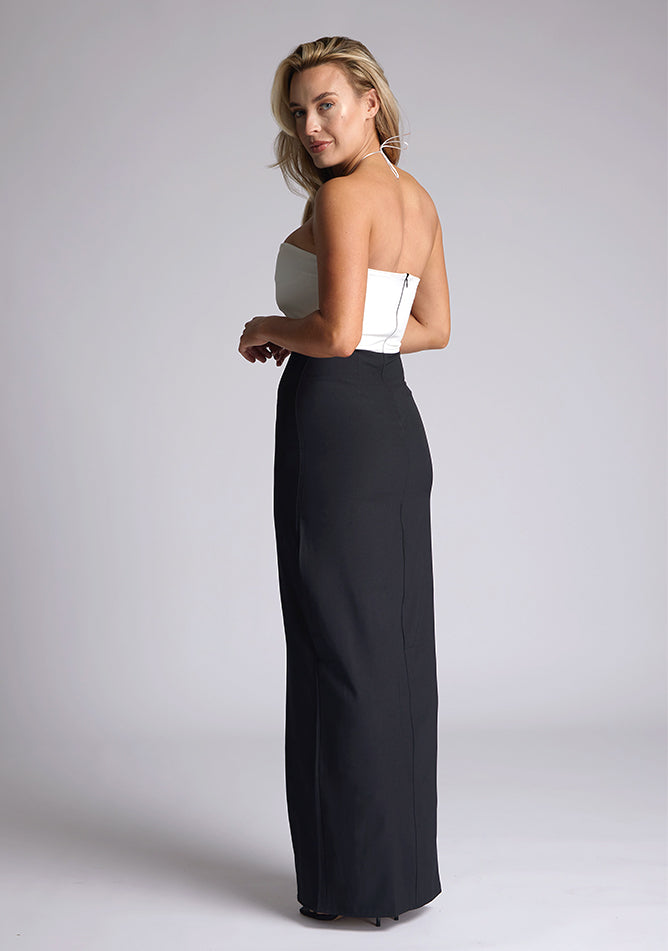 Quarter back image of a model wearing a black and white maxi dress, featuring a subtle sweetheart neckline, thin halter neck straps, a small keyhole cut-out at bust and a front skirt split in the middle. This maxi dress showcases an elegant ivory bust with a black maxi, the style featured is Vesper Heather Monochrome Maxi Dress.