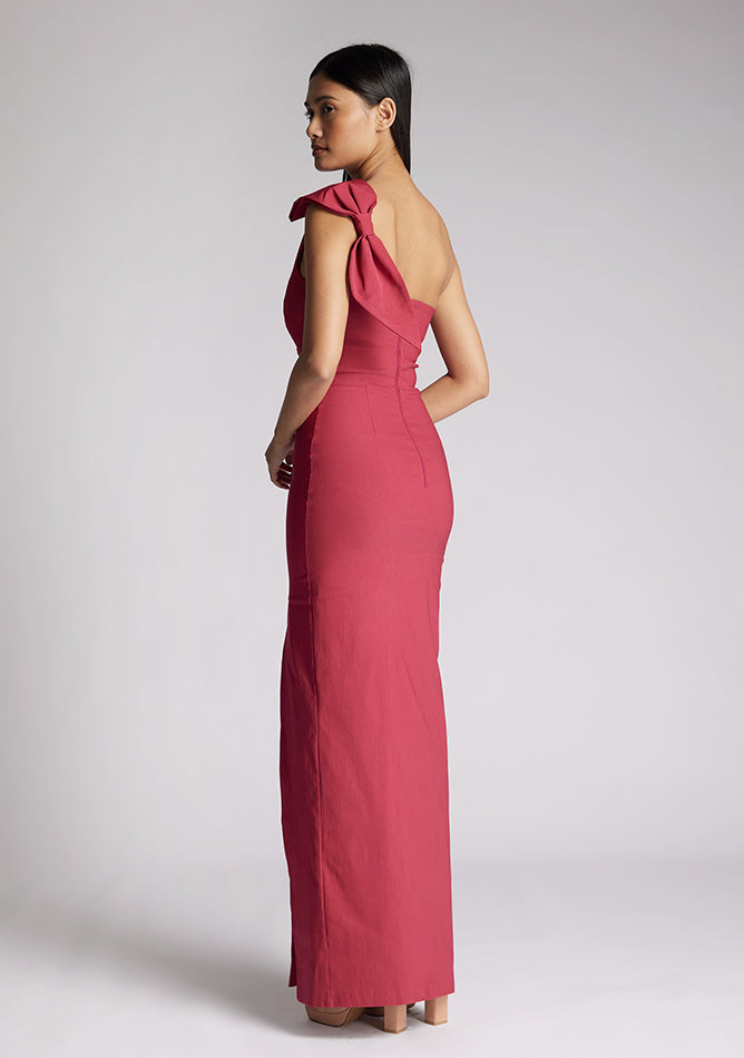 Back quarter image of a model wearing a raspberry maxi dress, featuring an asymmetric neckline, one shoulder with statement bow detail and a front skirt split. The dress featured is Vesper Keegan Raspberry One Shoulder Maxi Dress