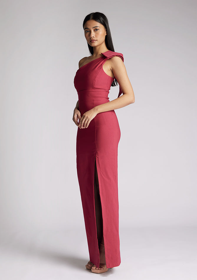 Front quarter image of a model wearing a raspberry maxi dress, featuring an asymmetric neckline, one shoulder with statement bow detail and a front skirt split. The dress featured is Vesper Keegan Raspberry One Shoulder Maxi Dress