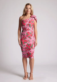 Front image of a model wearing a pink floral print midi dress, featuring an asymmetric neckline and one shoulder with a statement bow detail. The dress featured is the Vesper London Pink Floral One Shoulder Midi Dress.