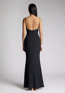 Back iimage of a model wearing a black maxi dress with a v-neckline, thin straps with twist front detail at bust, low back and subtle flair at hem. The design featured is the Vesper Marlowe Black Maxi Dress