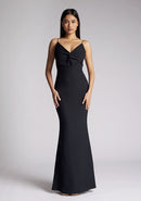 Front image of a model wearing a black maxi dress with a v-neckline, thin straps with twist front detail at bust, low back and subtle flair at hem. The design featured is the Vesper Marlowe Black Maxi Dress