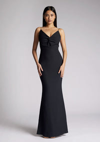 Front image of a model wearing a black maxi dress with a v-neckline, thin straps with twist front detail at bust, low back and subtle flair at hem. The design featured is the Vesper Marlowe Black Maxi Dress