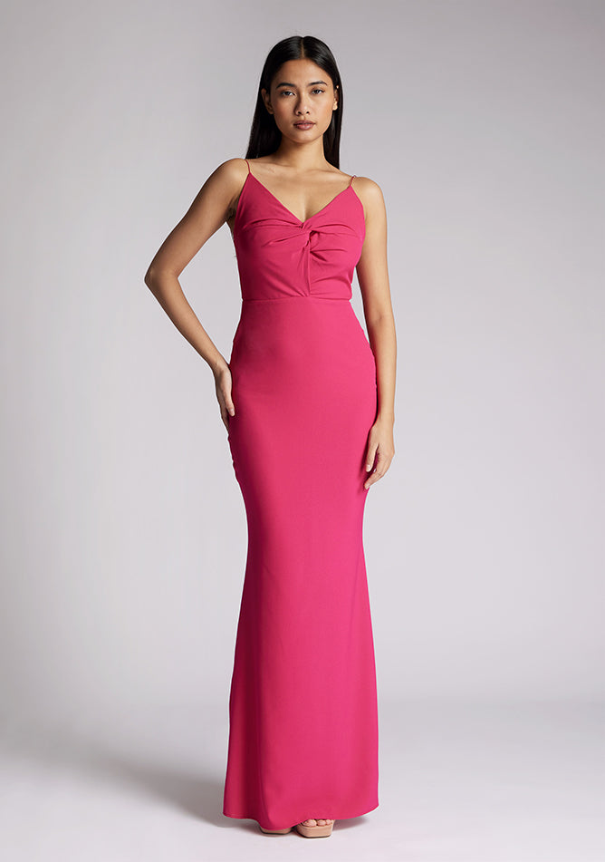 Front image of a model wearing a cerise pink maxi dress, featuring a v-neckline, thin straps with twist front detail at bust, low back and subtle flair at hem. The style featured is Vesper Marlowe Cerise Maxi Dress.