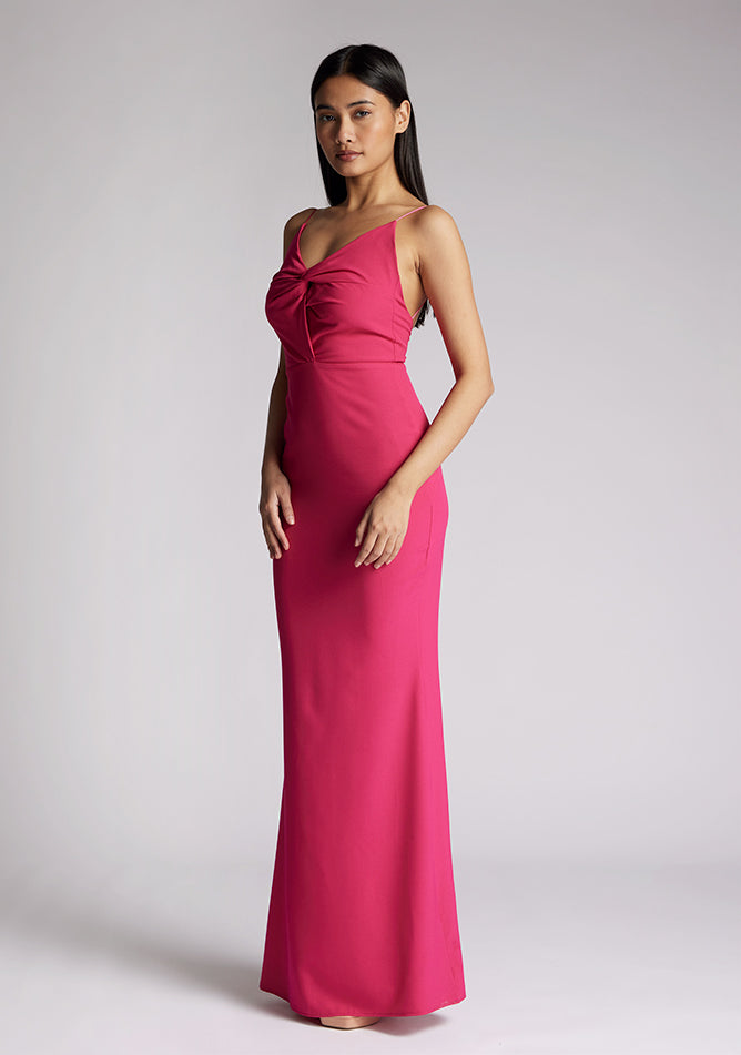 Quarter front image of a model wearing a cerise pink maxi dress, featuring a v-neckline, thin straps with twist front detail at bust, low back and subtle flair at hem. The style featured is Vesper Marlowe Cerise Maxi Dress.