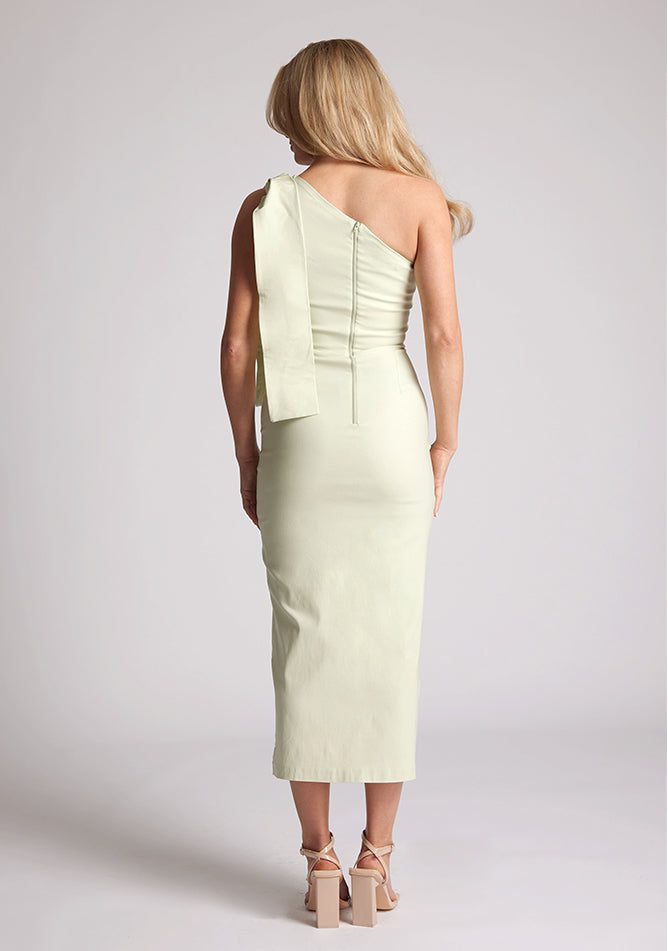 Back image of a model wearing a sage midaxi dress, featuring an asymmetric neckline with bow detail at shoulder, front skirt split and centre back invisible zip.The dress featured is the Vesper Odette Sage Midaxi Dress