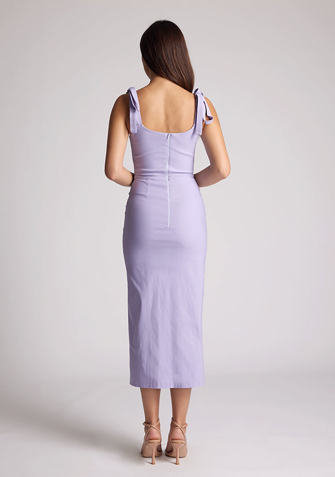 Back image of a model wearing a lilac midaxi dress, featuring tie up straps and a front skirt split. The dress featured is the Vesper Onyx lilac midaxi dress