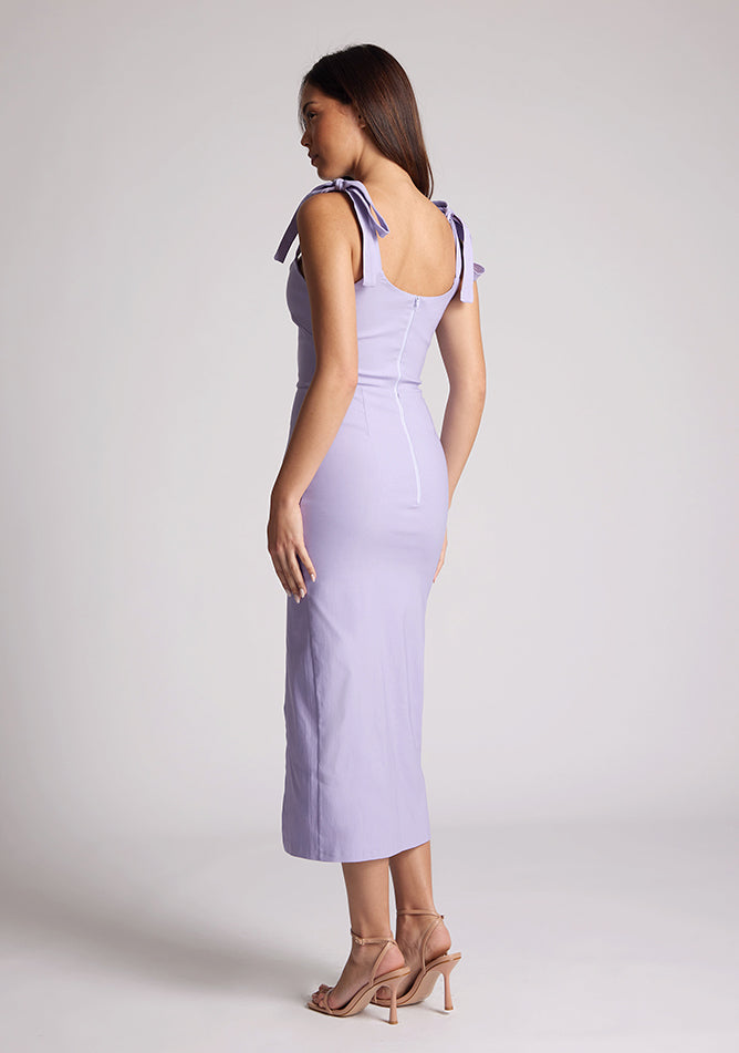 Back quarter image of a model wearing a lilac midaxi dress with a featuring scoop neckline, tie shoulder straps up, front skirt split and bodycon fit accentuates curves for finesse. The design featured is the Vesper Onyx Lilac Midaxi Dress
