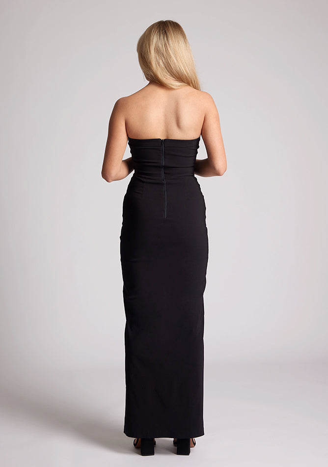 Back image of a model wearing a black maxi dress with a straight strapless neckline, body-hugging silhouette, front skirt split and invisible centre back zip. The design featured is the Vesper Raine Black Strapless Maxi Dress