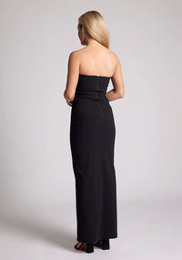 Back quarter image of a model wearing a black maxi dress with a straight strapless neckline, body-hugging silhouette, front skirt split and invisible centre back zip. The design featured is the Vesper Raine Black Strapless Maxi Dress