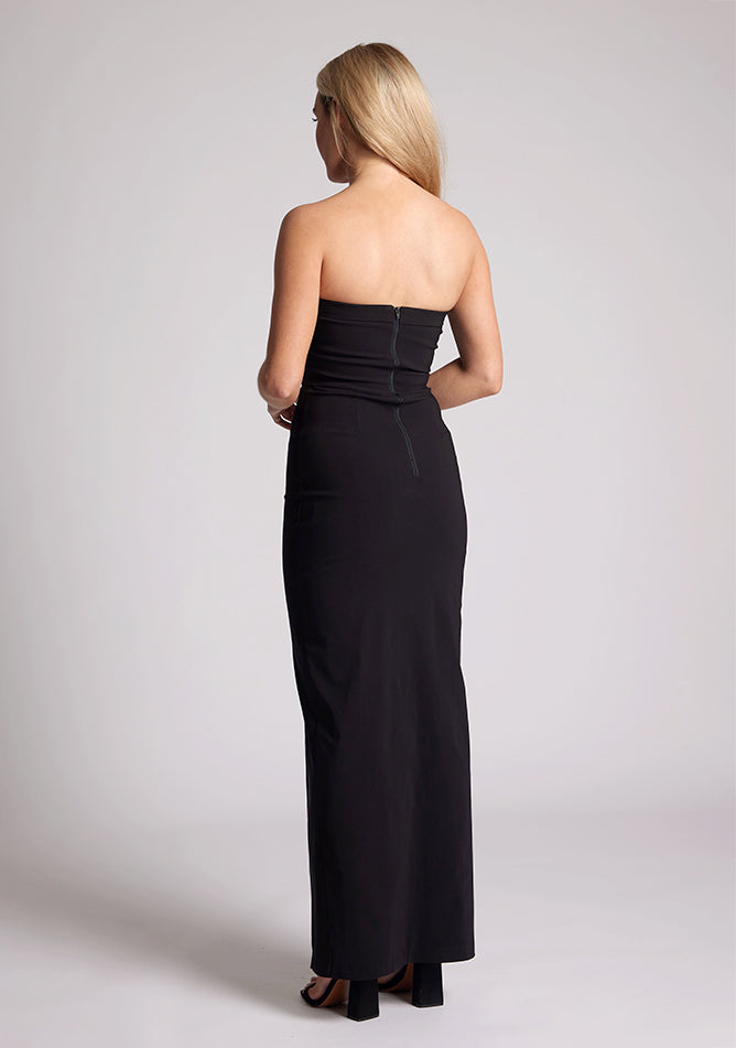 Back quarter image of a model wearing a black maxi dress with a straight strapless neckline, body-hugging silhouette, front skirt split and invisible centre back zip. The design featured is the Vesper Raine Black Strapless Maxi Dress