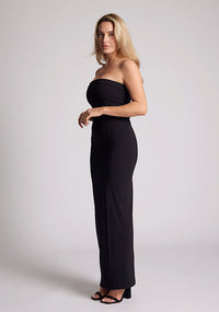 Front quarter image of a model wearing a black maxi dress with a straight strapless neckline, body-hugging silhouette, front skirt split and invisible centre back zip. The design featured is the Vesper Raine Black Strapless Maxi Dress