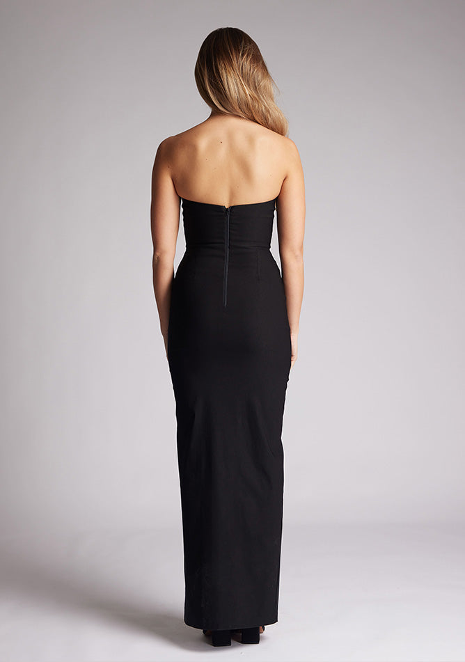 Back image of a blonde model wearing a black bodycon maxi dress with a  strapless elegance with a front skirt split for graceful movement and flattering slim fit enhances silhouette.  The design featured is the Vesper Remington Black Bodycon Maxi Dress
