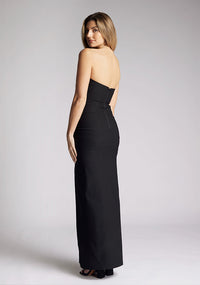 Back quarter image of a blonde model wearing a black bodycon maxi dress with a  strapless elegance with a front skirt split for graceful movement and flattering slim fit enhances silhouette.  The design featured is the Vesper Remington Black Bodycon Maxi Dress