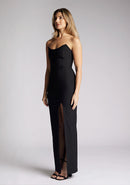 Front  quarter image of a blonde model wearing a black bodycon maxi dress with a  strapless elegance with a front skirt split for graceful movement and flattering slim fit enhances silhouette.  The design featured is the Vesper Remington Black Bodycon Maxi Dress