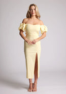 Front image of a blonde model wearing a yellow bardot midaxi dress with a subtle sweetheart neckline, statement puff sleeves, a front skirt split and lace-up detail at the back. The design featured is the Vesper River Yellow Bardot Midaxi Dress