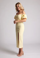 Front quarter image of a blonde model wearing a yellow bardot midaxi dress with a subtle sweetheart neckline, statement puff sleeves, a front skirt split and lace-up detail at the back. The design featured is the Vesper River Yellow Bardot Midaxi Dress