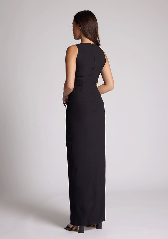Back quarter image of a model wearing a black maxi dress, featuring a round neckline, side bodice cut-out, side waist pleating detail and front skirt split. The dress featured is the Vesper Rowan Black Maxi Cut Out Dress