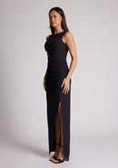 Front quarter image of a model wearing a black maxi dress, featuring a round neckline, side bodice cut-out, side waist pleating detail and front skirt split. The dress featured is the Vesper Rowan Black Maxi Cut Out Dress