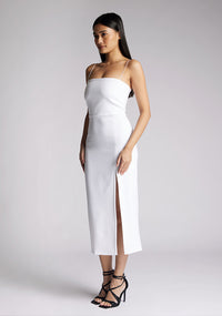Front image of a model wearing a white midaxi dress with a minimalistic strap details, front skirt split and centre back zip. The design featured is the Vesper Tate White Midaxi Dres