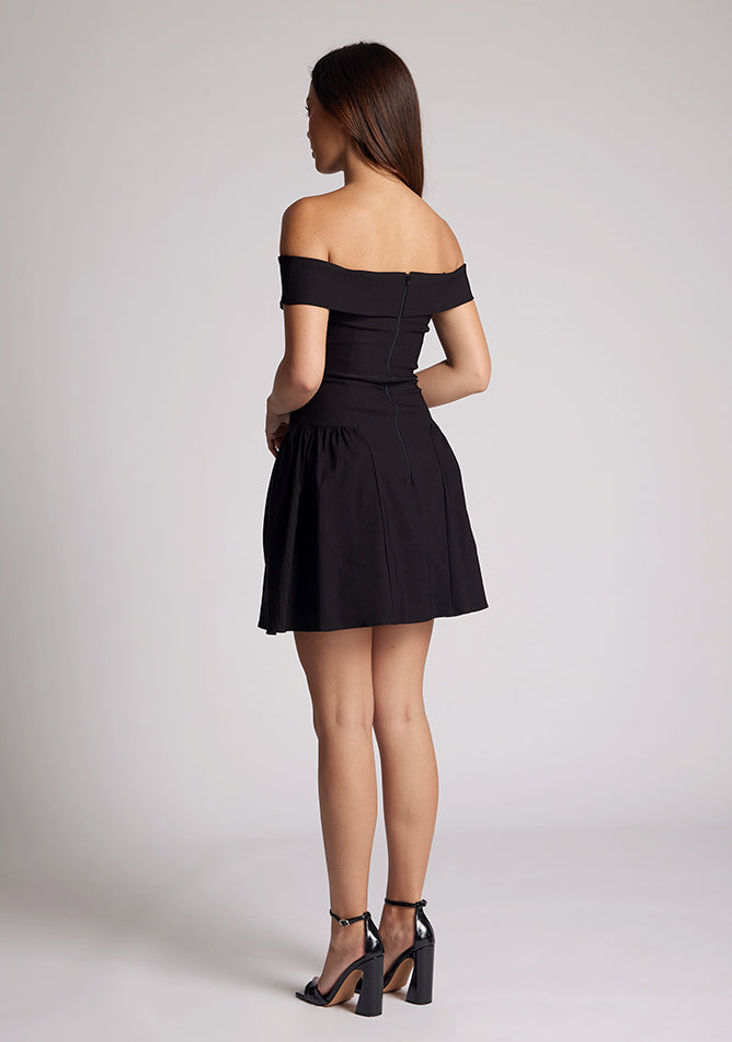 Back quarter image of a model wearing a black mini dress with a straight neckline, with an off-shoulder design, bold bardot brand across the arm and neckline and fitted bodice with A-line skirt and centre back invisible zip. The design featured is the Vesper Una Black Mini Dress