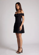 Front quarter image of a model wearing a black mini dress with a straight neckline, with an off-shoulder design, bold bardot brand across the arm and neckline and fitted bodice with A-line skirt and centre back invisible zip. The design featured is the Vesper Una Black Mini Dress.