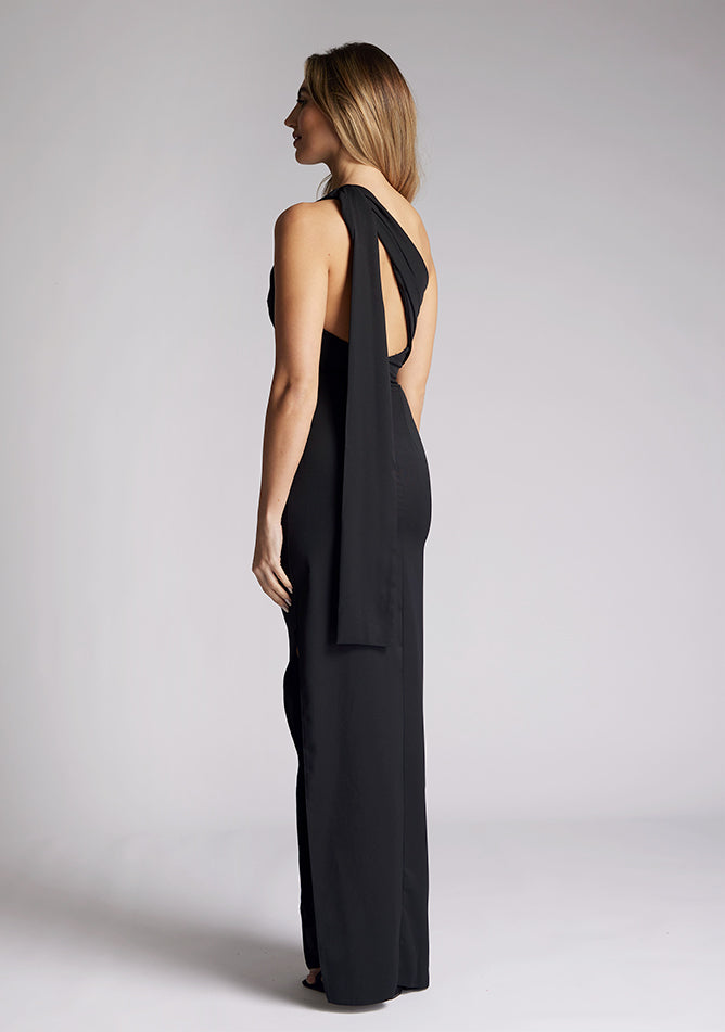 Quarter back image of a model wearing a black maxi dress, featuring a one shoulder design with draped fabric at shoulder, and a statement asymmetric back with cut-out details and front skirt split.  The dress featured is the Vesper Drew black maxi dress