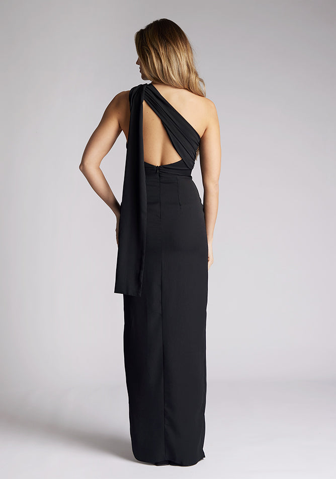 Back image of a model wearing a black maxi dress, featuring a one shoulder design with draped fabric at shoulder, and a statement asymmetric back with cut-out details and front skirt split.  The dress featured is the Vesper Drew black maxi dress
