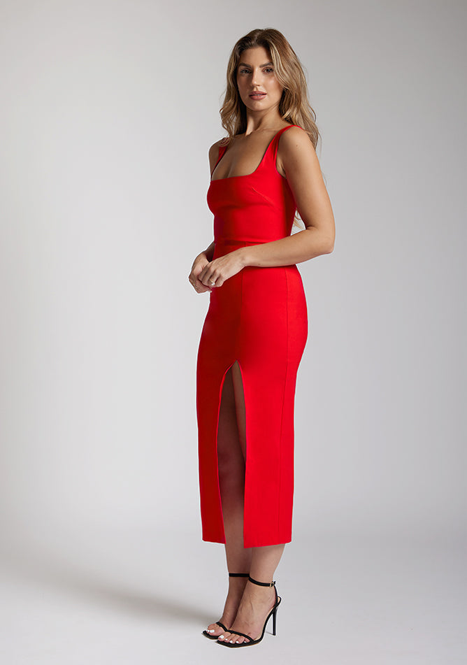 Front quarter image of a blonde model wearing a red bodycon midaxi dress with a square neckline with wide straps and a front skirt split. The design featured is the Vesper Abbie Red Bodycon Midaxi Dress