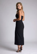 Back quarter image of the model wearing a black bodycon midaxi dress with a straight neckline, cross strap detail, and front skirt split. The design featured is the Vesper Abigail Black Cross Strap Midaxi Dress