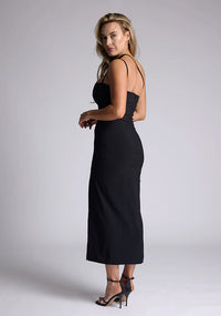 Back quarter image of the model wearing a black bodycon midaxi dress with a straight neckline, cross strap detail, and front skirt split. The design featured is the Vesper Abigail Black Cross Strap Midaxi Dress