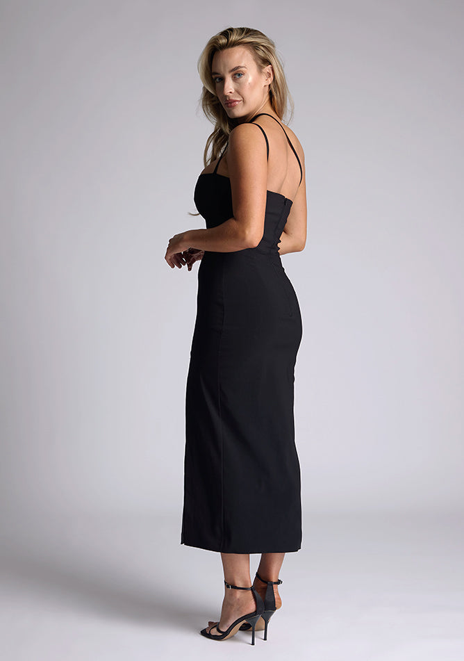 Back quarter image of the model wearing a black bodycon midaxi dress with a straight neckline, cross strap detail, and front skirt split. The design featured is the Vesper Abigail Black Cross Strap Midaxi Dress
