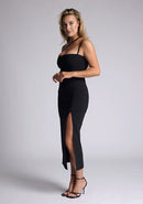 Front quarter image of the model wearing a black bodycon midaxi dress with a straight neckline, cross strap detail, and front skirt split. The design featured is the Vesper Abigail Black Cross Strap Midaxi Dress