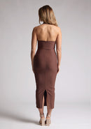 Back image of a blonde model wearing a chocolate midaxi dress with a halter neckline, sleeveless, and a back skirt split. The design features is the Vesper Asher Chocolate Halter Neck Midaxi Dress