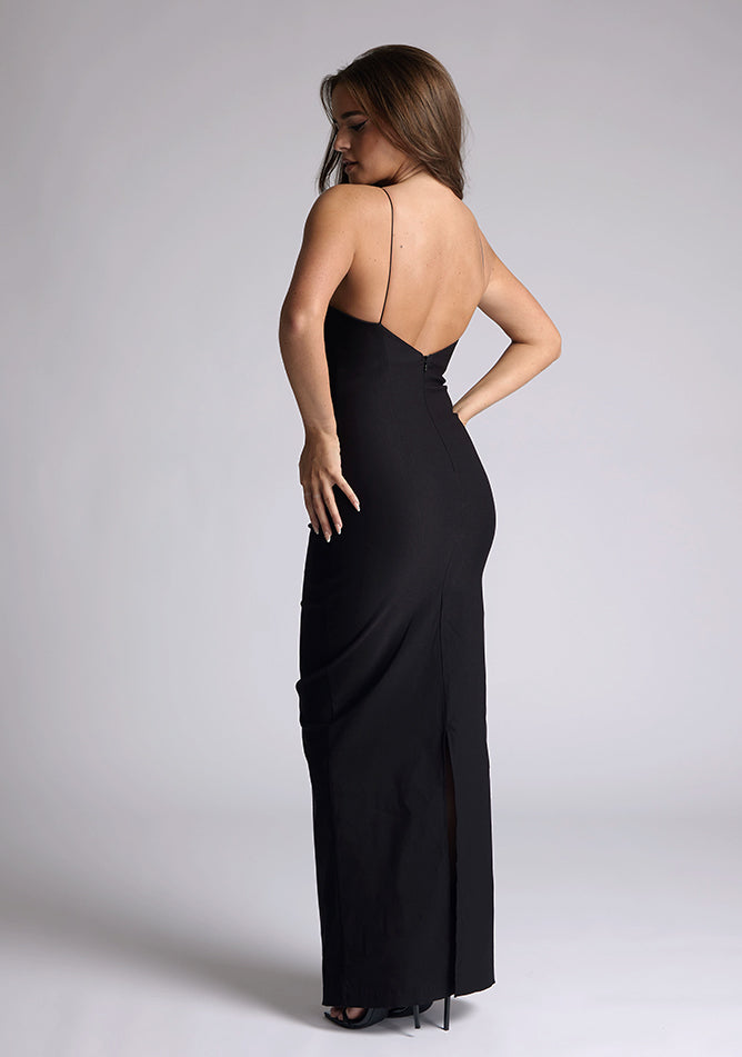 Back quarter image of a model wearing a black maxi dress with a plunge neckline and bow detailing. The design featured is the Vesper Bellini Black Bow Maxi Dress