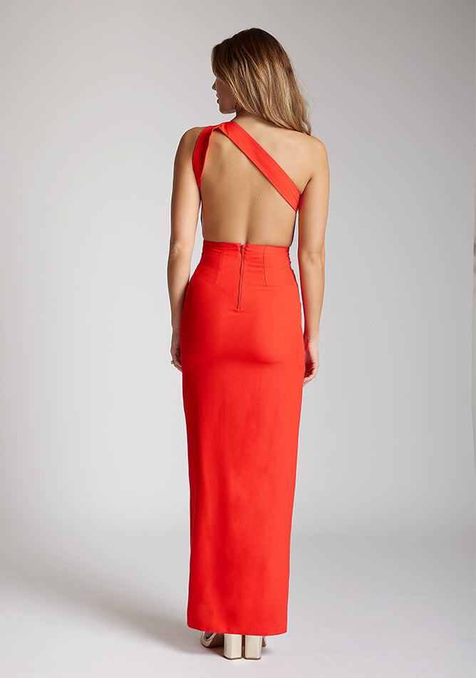 Back image of a model wearing a red maxi dress with a asymmetric neckline, with an open back and an asymmetric statement strap across the back and front skirt split. The designe featured is Vesper Deva Red Maxi Dress