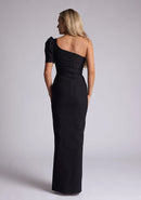 Back image of a blonde model wearing a black maxi dress with a one puff sleeve design adds a touch of sophistication, while the front skirt split enhances movement and grace. The design features a Vesper Evangelical Black One Sleeve Maxi Dress