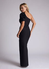 Back quarter image of a blonde model wearing a black maxi dress with a one puff sleeve design adds a touch of sophistication, while the front skirt split enhances movement and grace. The design features a Vesper Evangelical Black One Sleeve Maxi Dress
