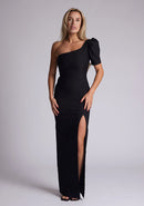 Front image of a blonde model wearing a black maxi dress with a one puff sleeve design adds a touch of sophistication, while the front skirt split enhances movement and grace. The design features a Vesper Evangelical Black One Sleeve Maxi Dress