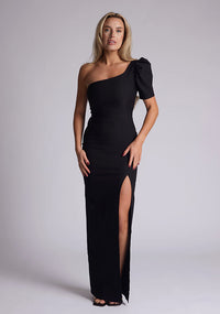 Front image of a blonde model wearing a black maxi dress with a one puff sleeve design adds a touch of sophistication, while the front skirt split enhances movement and grace. The design features a Vesper Evangelical Black One Sleeve Maxi Dress
