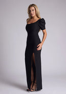 Front quarter image of a blonde model wearing a black maxi dress with a one puff sleeve design adds a touch of sophistication, while the front skirt split enhances movement and grace. The design features a Vesper Evangelical Black One Sleeve Maxi Dress