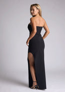 Back quarter image of blonde model wearing a black maxi dress with a  features a sweetheart neckline, centre back zip and a side skirt split. The design featured is the Vesper Larissa Black Bodycon Maxi Dress