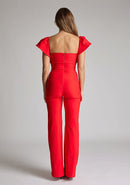 Back image of blonde model wearing a Red Jumpsuit with a  ruffled sleeves adding a touch of playfulness, a charming sweetheart neckline, and a convenient back zip, this outfit exudes timeless sophistication. The wide-leg design offers comfort and style, a design features Vesper Leigh Red Wide Leg Jumpsuit