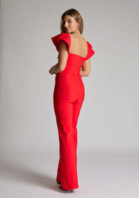 Back quarter image of blonde model wearing a Red Jumpsuit with a  ruffled sleeves adding a touch of playfulness, a charming sweetheart neckline, and a convenient back zip, this outfit exudes timeless sophistication. The wide-leg design offers comfort and style, a design features Vesper Leigh Red Wide Leg Jumpsuit