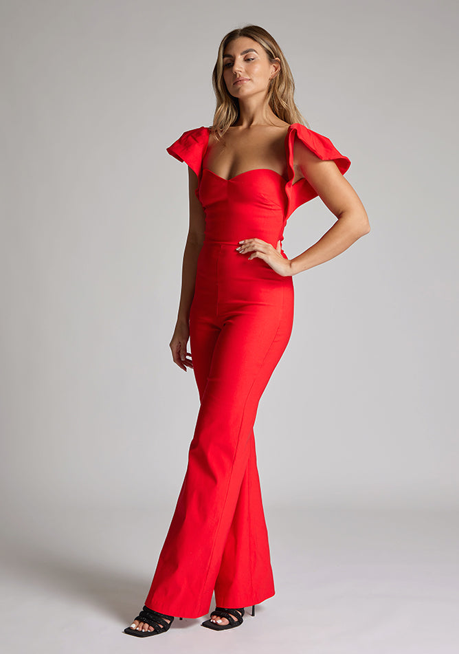 Front quarter image of blonde model wearing a Red Jumpsuit with a  ruffled sleeves adding a touch of playfulness, a charming sweetheart neckline, and a convenient back zip, this outfit exudes timeless sophistication. The wide-leg design offers comfort and style, a design features Vesper Leigh Red Wide Leg Jumpsuit