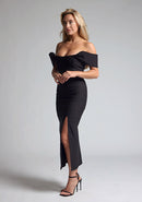 Front quarter image of blonde model wearing a Black Midaxi Dress with a off-shoulder design, a flattering body-con sillhoutte, and a front skirt split,. The design features is the Vesper Marriot Black Bardot Midaxi Dress