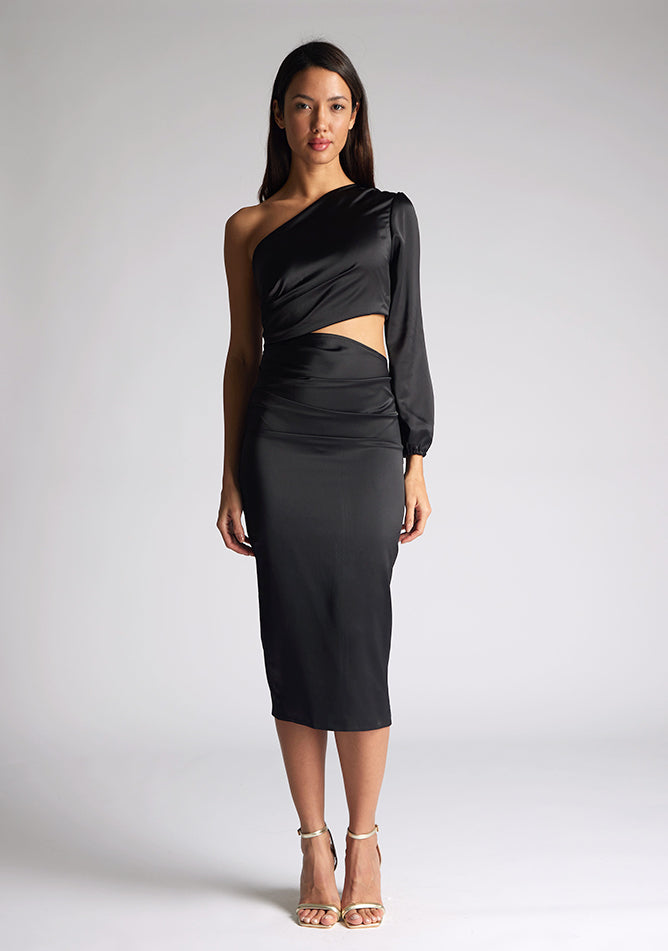 Front image of a model wearing an black midi dress, featuring an asymmetric neckline with one long sleeve, with a side waist cut-out detail.The dress featured is the Vesper Niccola Black Satin Midi Dress