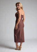 Back quarter image of a blonde model wearing a Chocolate Midaxi Dress with a daring plunge neckline, delicate thin straps, a flattering bodycon fit and a faux wrap skirt split adds a touch of sophistication to the overall design. The design features the Vesper Oaklynn Chocolate Midaxi Dress
