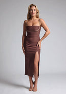Front image of a blonde model wearing a Chocolate Midaxi Dress with a daring plunge neckline, delicate thin straps, a flattering bodycon fit and a faux wrap skirt split adds a touch of sophistication to the overall design. The design features the Vesper Oaklynn Chocolate Midaxi Dress
