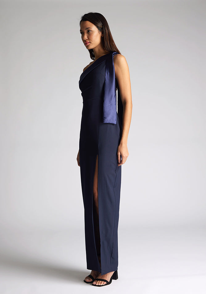 Front quarter image of a model wearing a  One Shoulder Navy Maxi Dress with a  one shoulder contrast satin band and bow detail with a front skirt split. The design features Vesper Olympia One Shoulder Navy Maxi Dress